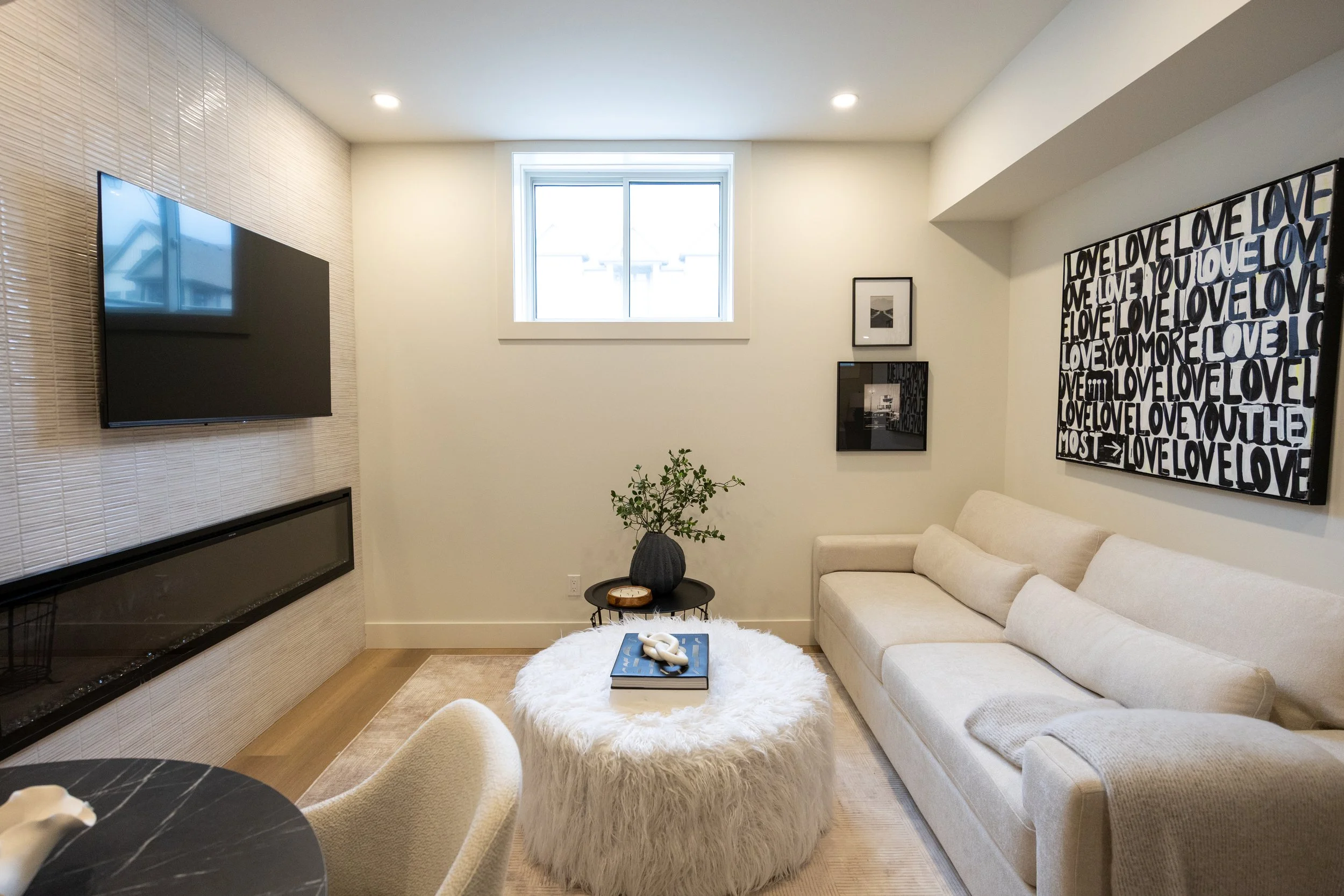 A cozy living room with a beige sofa, a white fluffy ottoman, a small round black side table with a plant, and wall artboards with words like "LOVE" and "YOU". There is a window with white trim and a wall-mounted flat-screen TV.