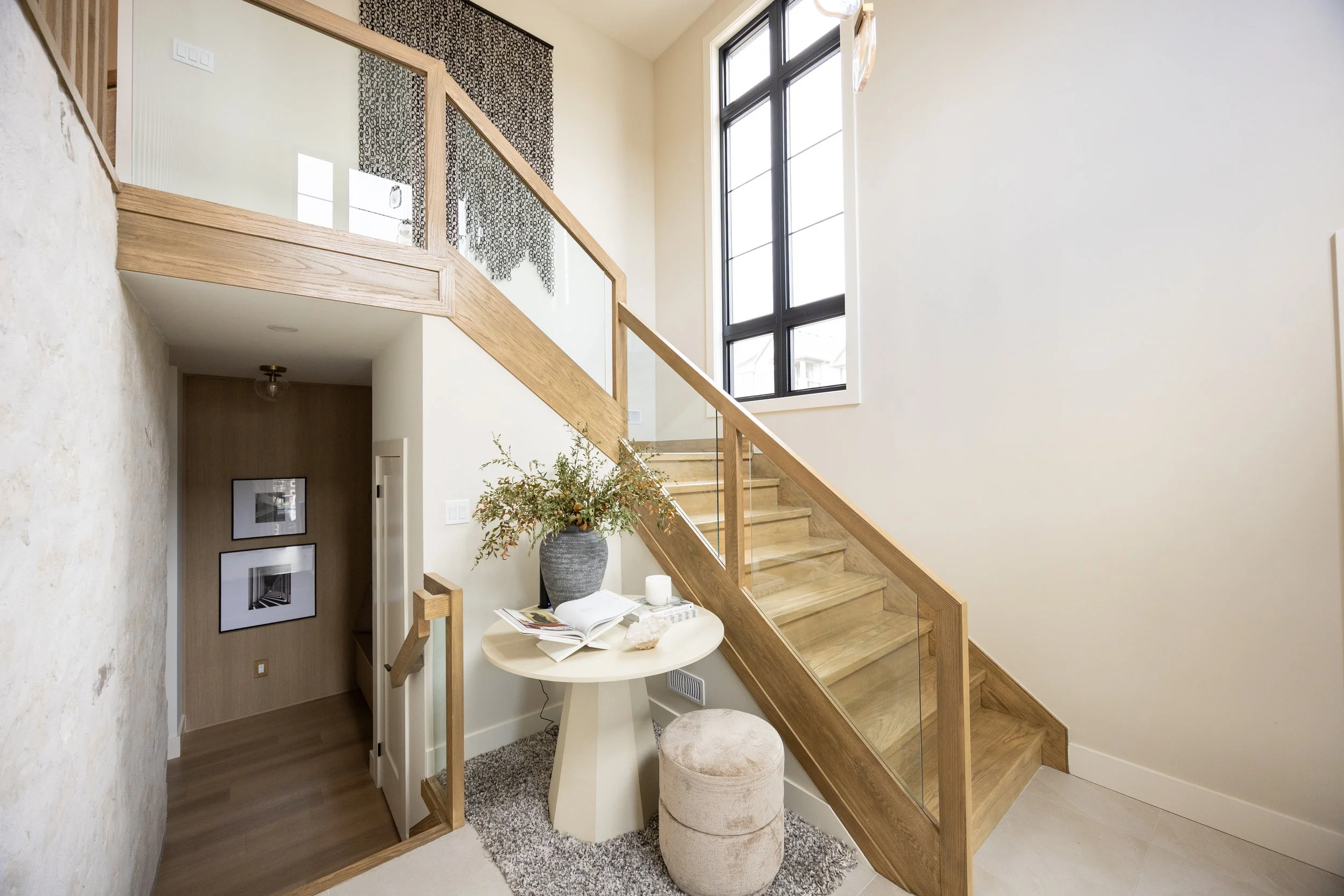Interior view of a staircase with wooden steps and glass railing, a large window letting in natural light, a small table with magazines and a potted plant, and wall art in the background.