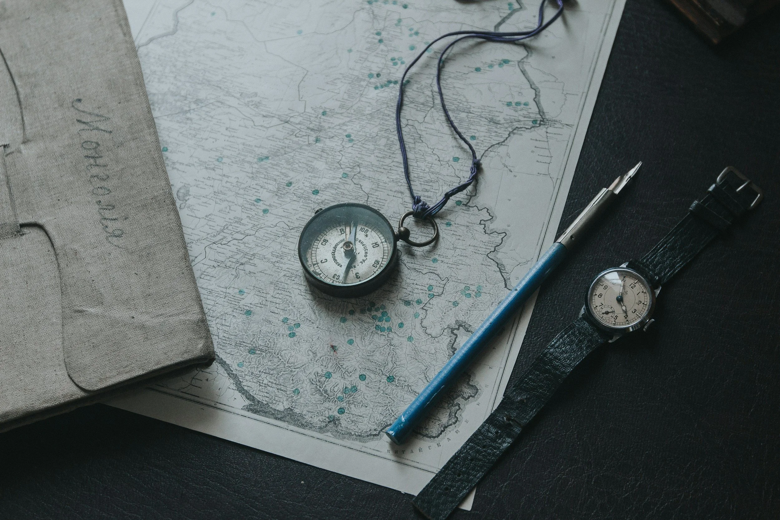 A vintage map with a compass, pocket watch, pen, and a fabric-covered book or journal on a dark surface.