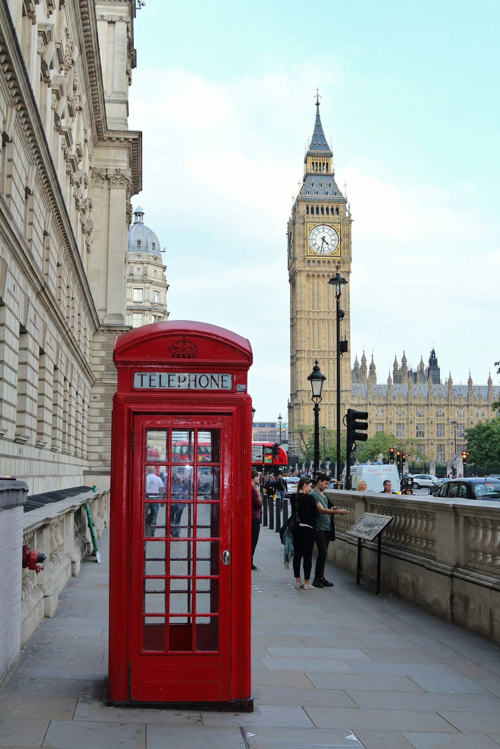A red British telephone booth on a sidewalk in London with Big Ben clock tower and historic buildings in the background, and people walking by.