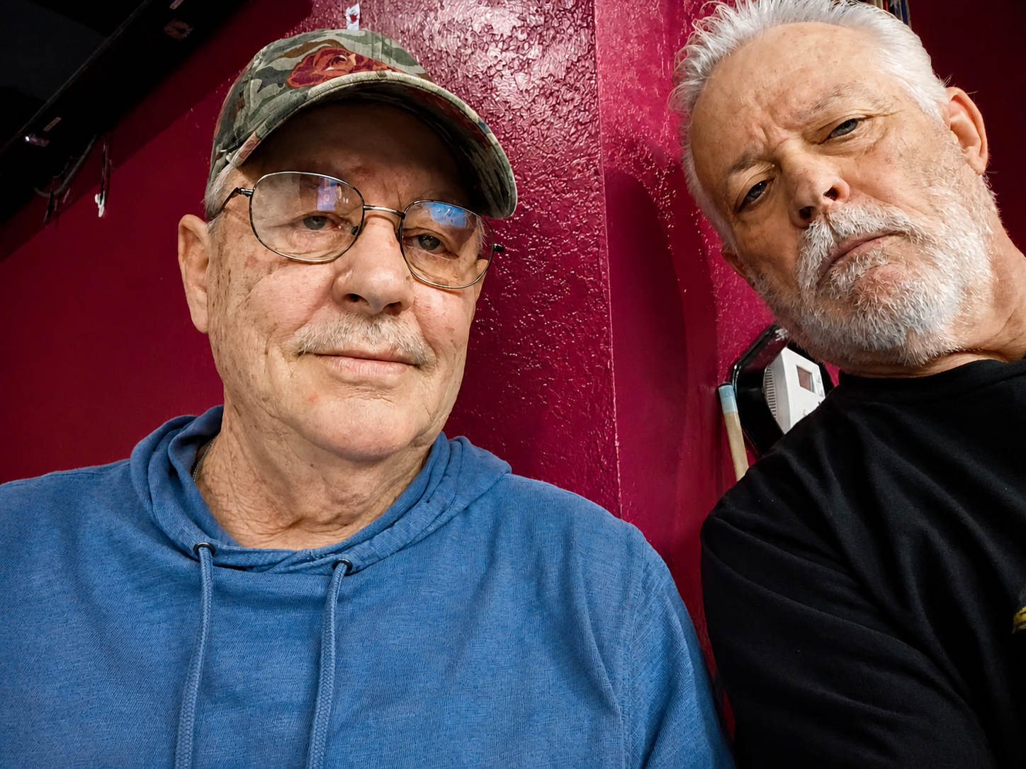 Two elderly men with white hair and beards sitting close together against a red textured wall. The man on the left is wearing glasses, a gray cap with a floral pattern, and a blue hoodie. The man on the right is wearing a black shirt. Both are lookin