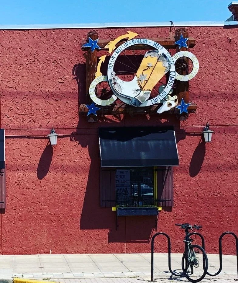 A sign created by Phyllis J Garibay-Coon for So Long Saloon in Manhattan, KS featuring at cowboy boot and wagon wheels. 