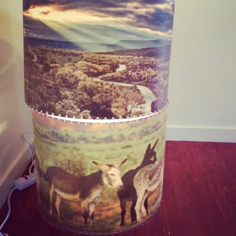 A cylindrical lampshade with landscape and animal scenes, including a river, mountains, trees, and two donkeys in a pastoral setting. Photo by Rod Mikinski, lampshades created by Phillis J Garibay-Coon.