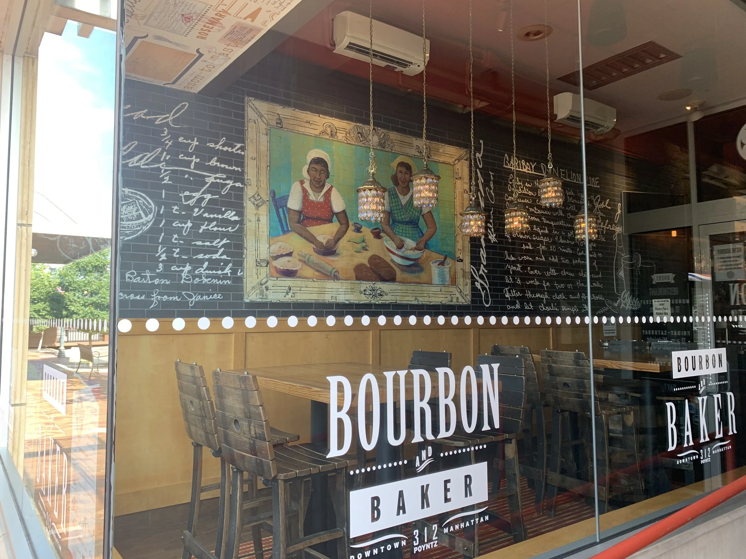 View inside a Bourbon & Baker in Manhattan, KS featuring a painting of two women baking and hanging pendant lights.