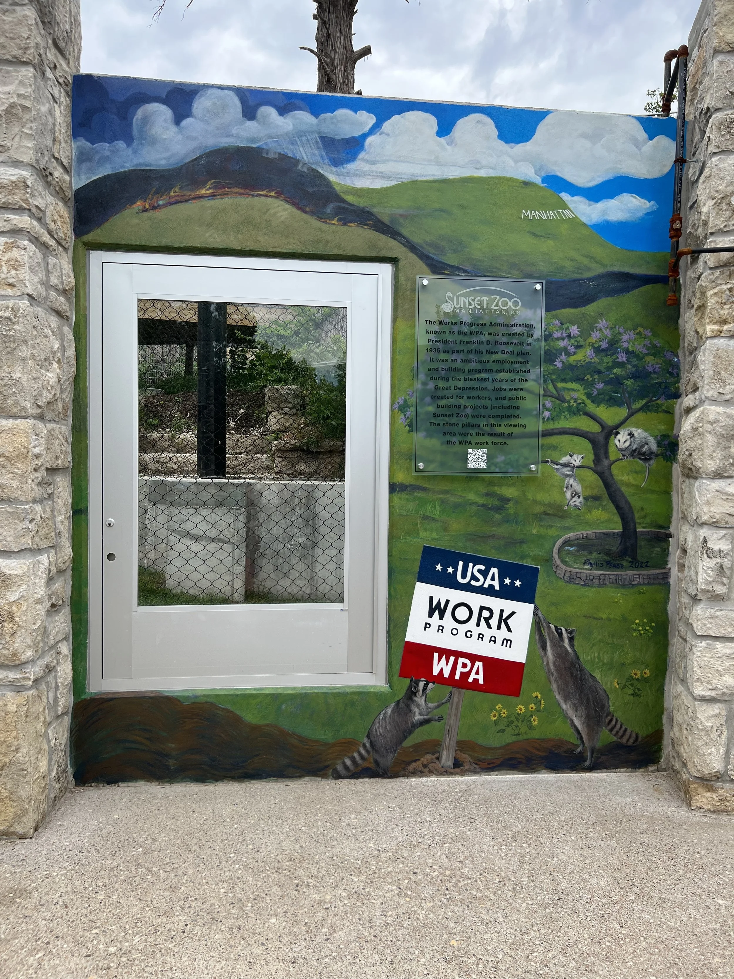 Colorful mural at Sunset Zoo depicting rolling hills, clouds, trees, and raccoons, with a sign in front of the mural about the zoo's history and a WPA Work Program sign.