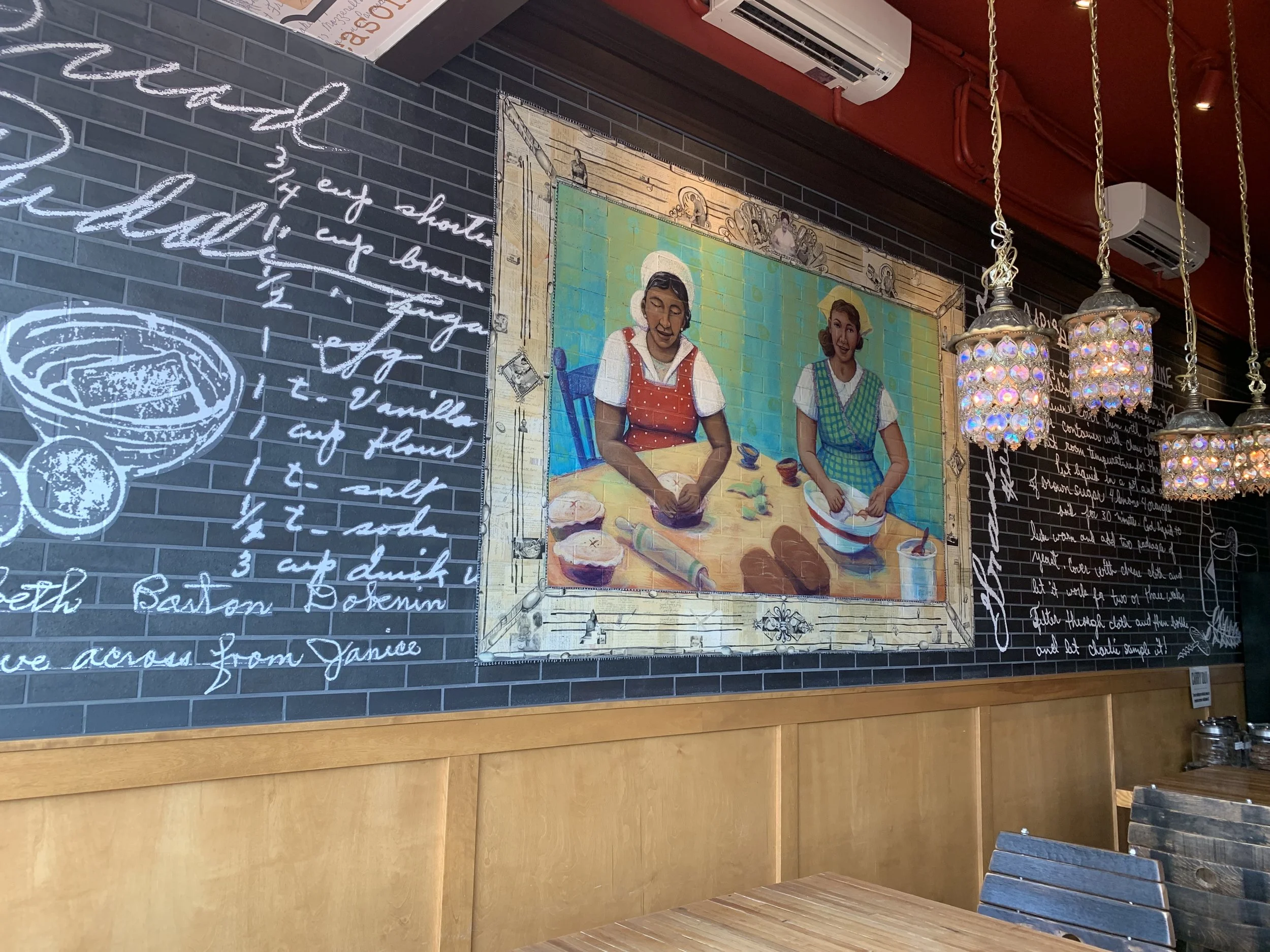 View inside a Bourbon & Baker in Manhattan, KS featuring a painting of two women baking and hanging pendant lights.