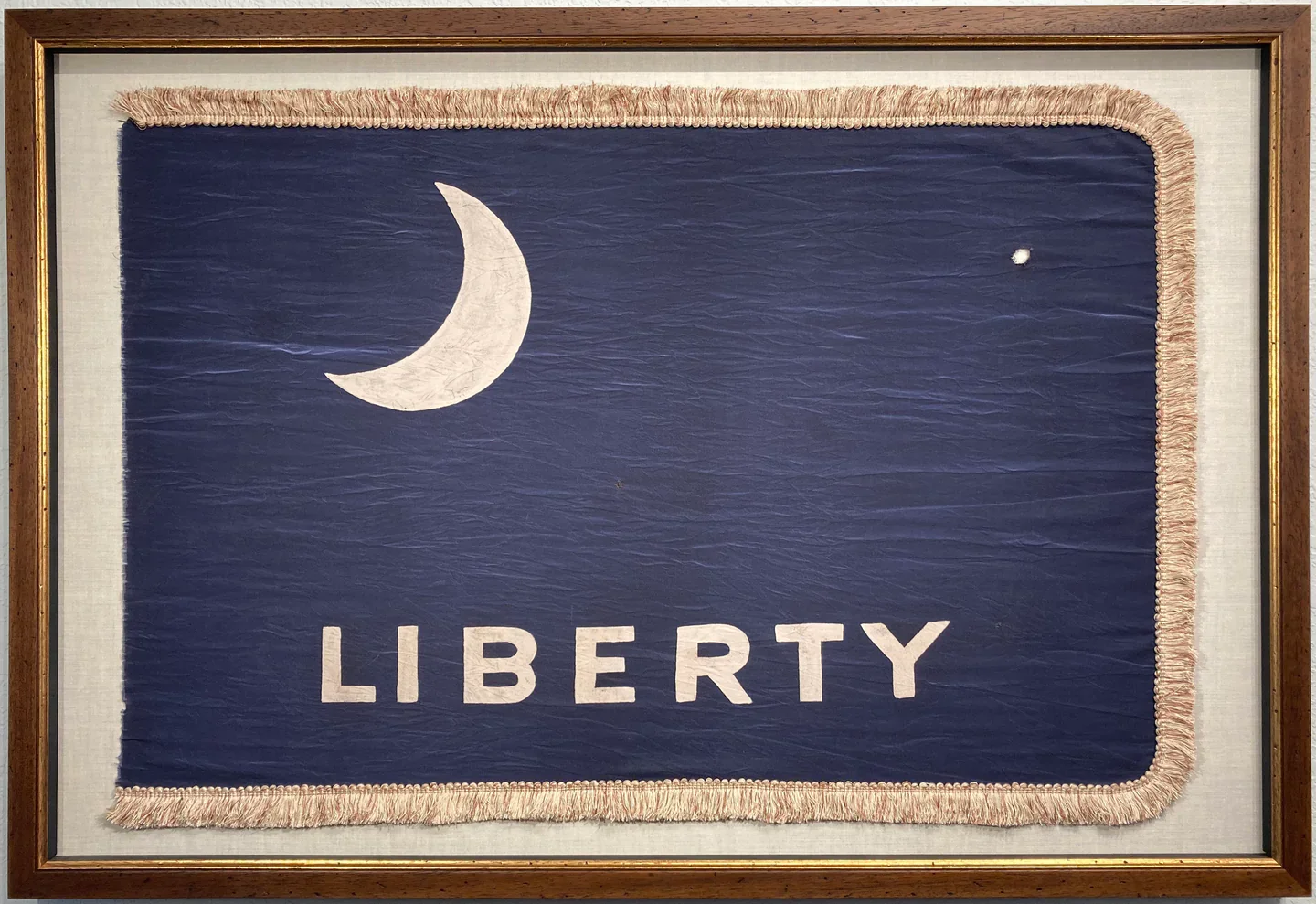 A framed fabric flag with a blue background, a white crescent moon, a small white star, and the word "LIBERTY" written in white letters.