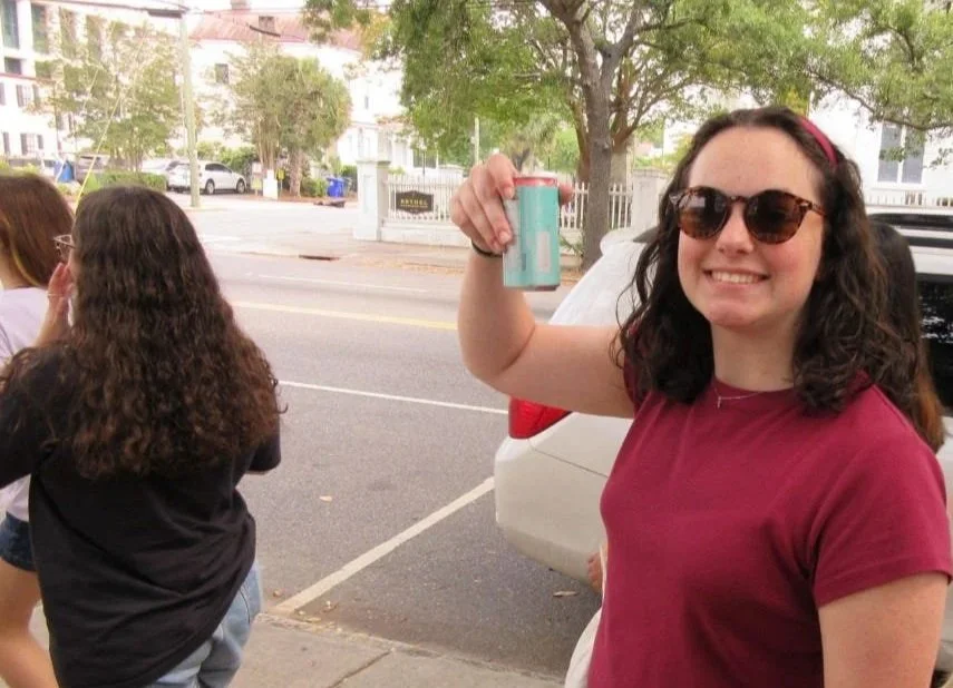 A young woman with curly dark hair, wearing sunglasses and a maroon shirt, smiling and holding a beverage can in her right hand. She is standing outdoors on a sidewalk near parked cars, with trees and other people in the background.