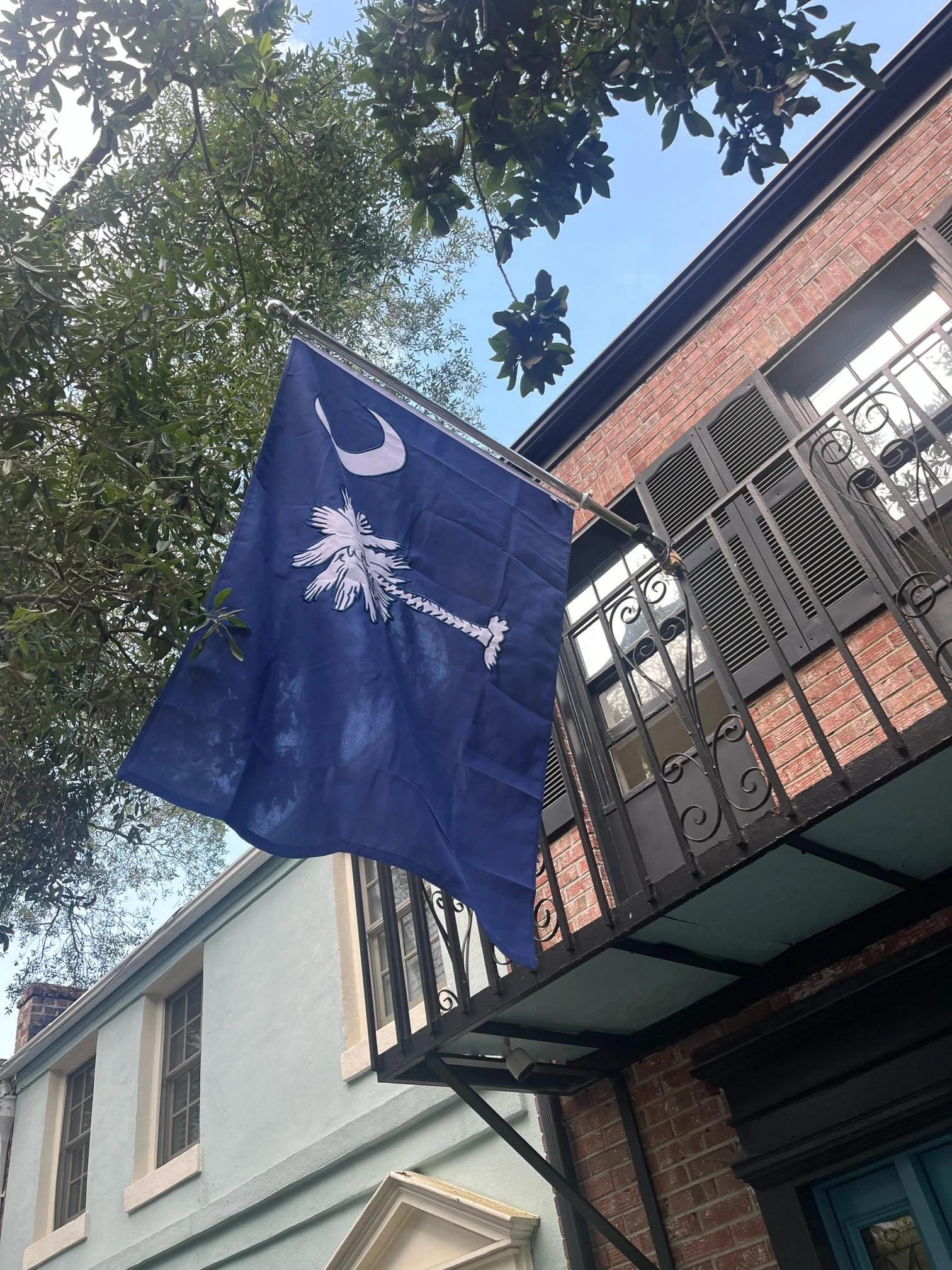 A blue flag with a white bird and a crescent moon is hanging from a black metal balcony railing on a brick building. Green trees and a blue sky are visible in the background.