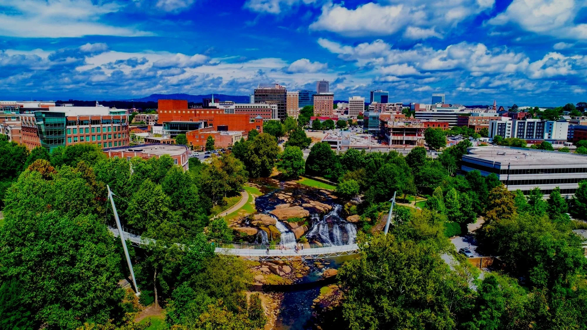 A city skyline with tall buildings, green trees, a waterfall, and a white suspension bridge under a partly cloudy blue sky.