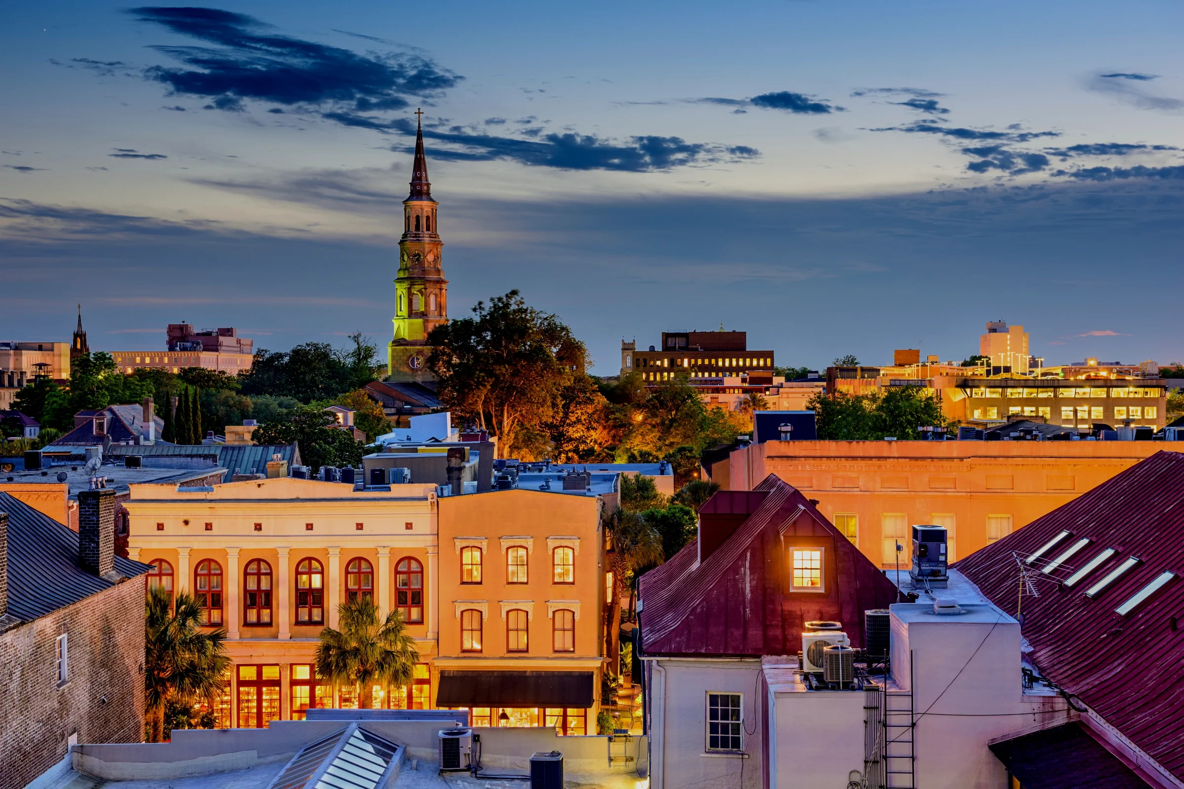 Cityscape at dusk showing buildings, trees, a church steeple, and a sky with clouds.