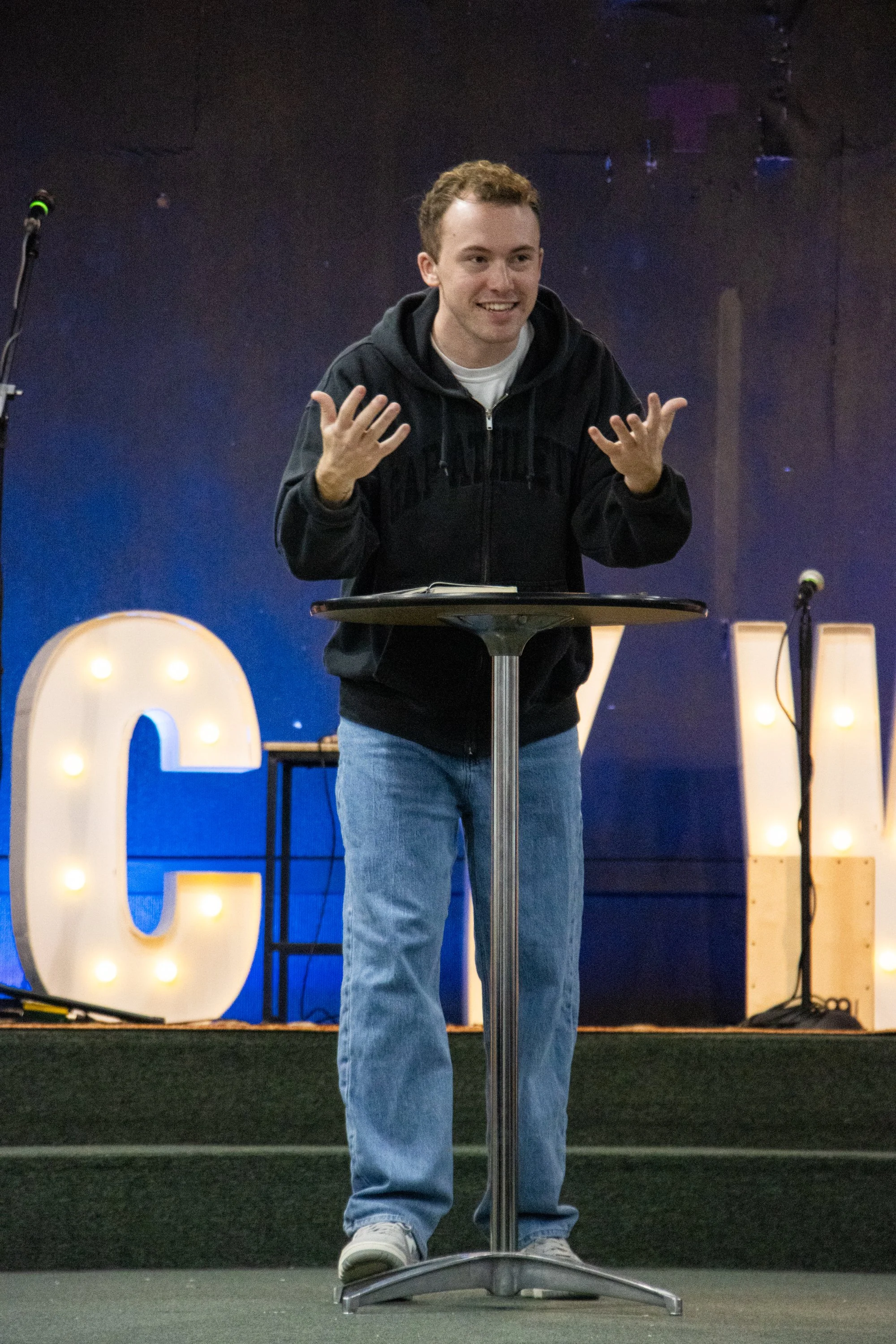 A young man with short, curly hair standing behind a round table, smiling, and gesturing with his hands. He is wearing a black hoodie and light blue jeans. Behind him are large illuminated letter signs spelling a word, with a dark, textured background and two microphones on stands.