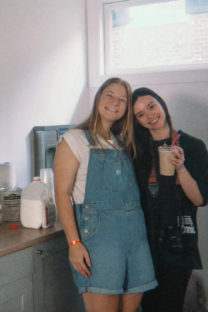 Two smiling women standing in a kitchen next to a counter. One woman is wearing denim overalls and a white t-shirt, and the other is holding a coffee cup and has a camera hanging around her neck. There are bottles and paper towels on the counter, and a window with a brick wall visible outside.