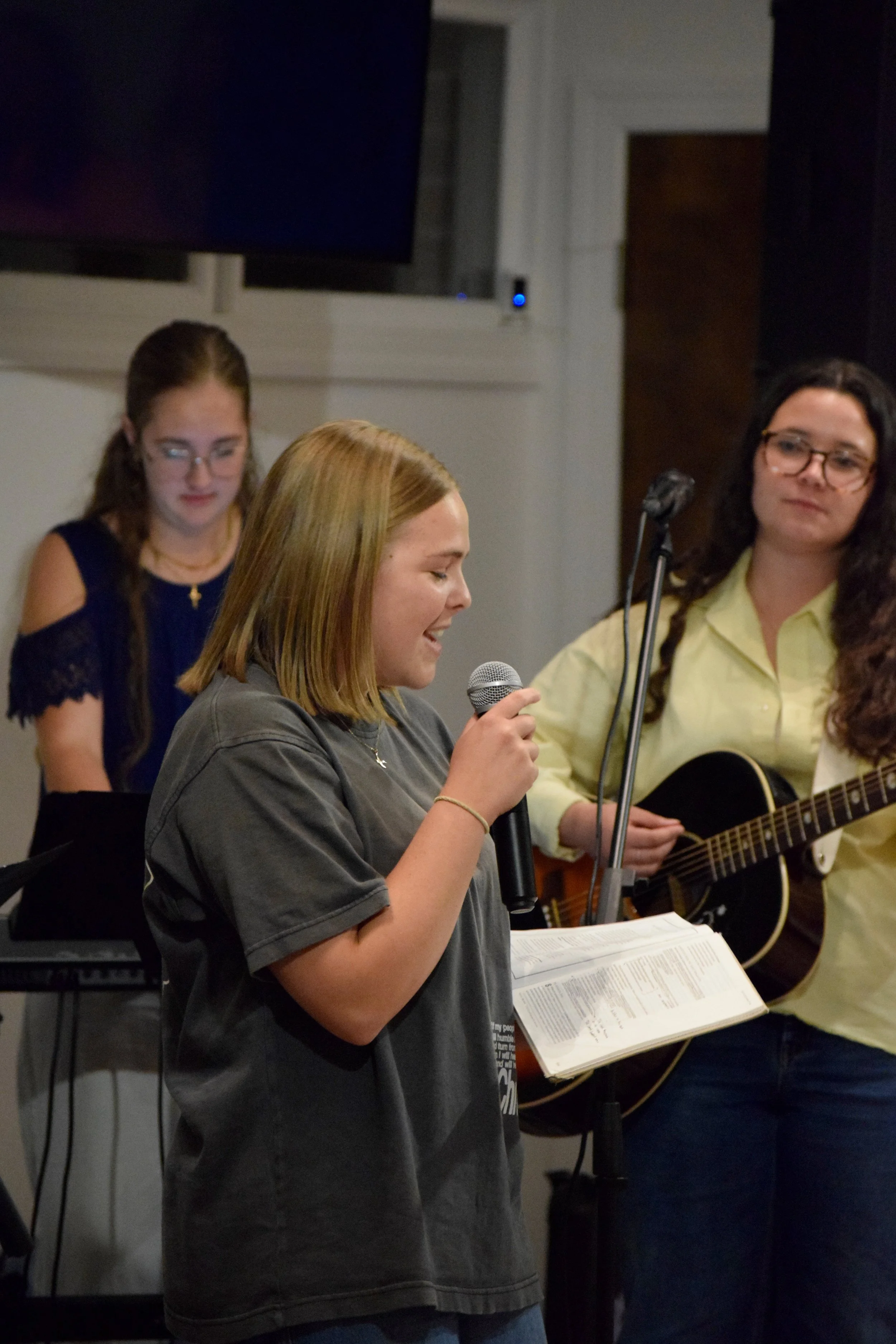 A group of young women performing music and singing; one is holding a microphone and a music sheet, another is playing a guitar.