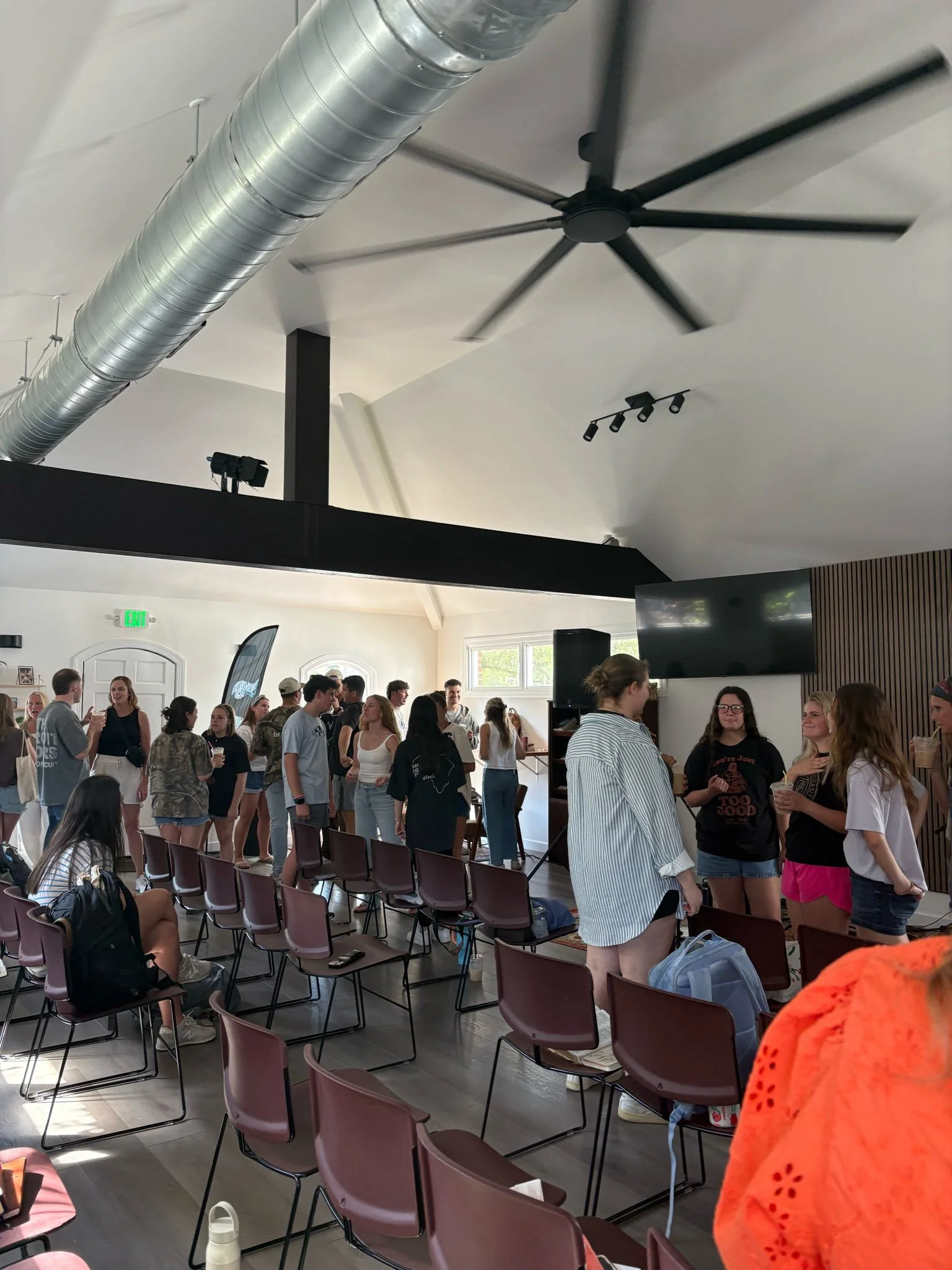 A large group of people gathered in a spacious room with high ceilings, light-colored walls, and a ceiling fan. Some people are standing, chatting, and others are seated on chairs, with many holding cups. The room has modern lighting and large windows allowing natural light inside.