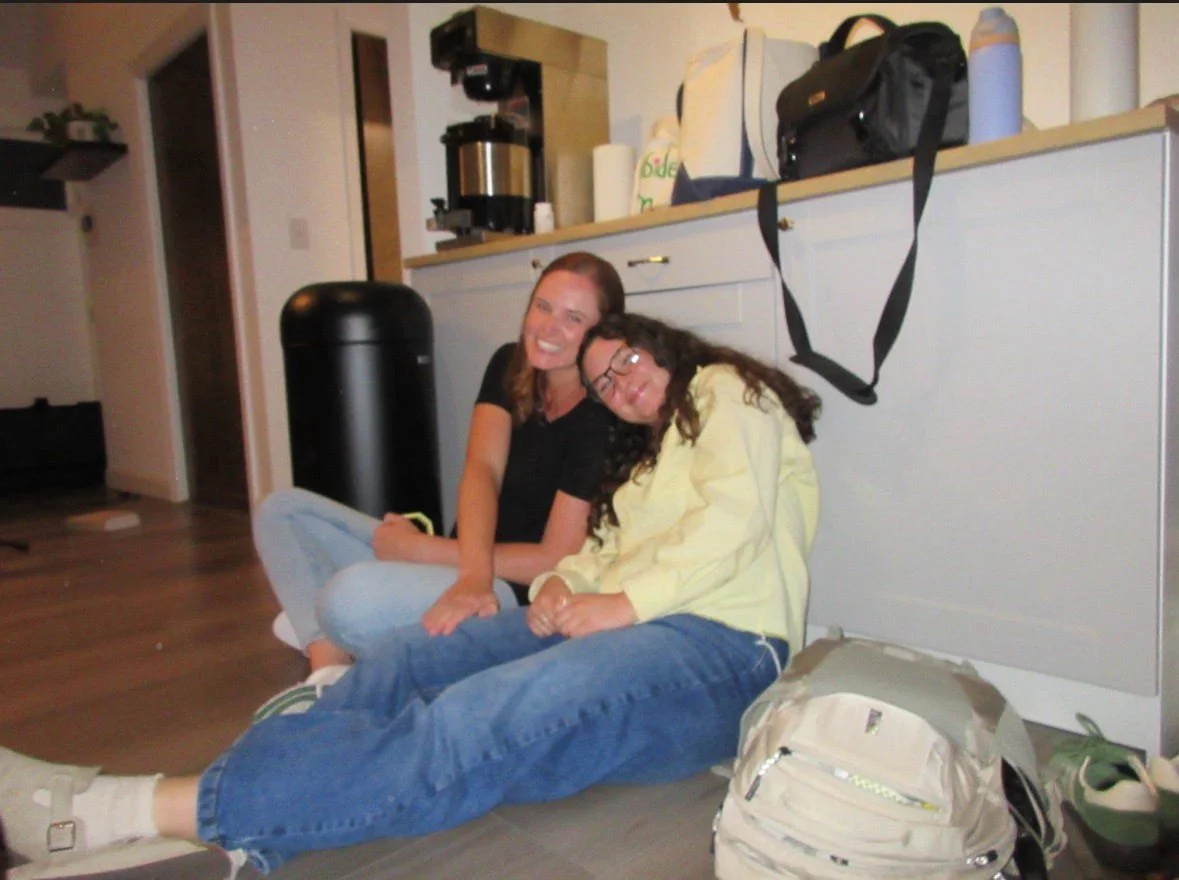 Two women sitting on the floor and smiling, with one woman leaning her head on the other's shoulder. They are in a room with a kitchen counter behind them, which has bags, a water bottle, and a black strap hanging from the counter. There's a large black trash can, a coffee maker, and a doorway visible in the background.