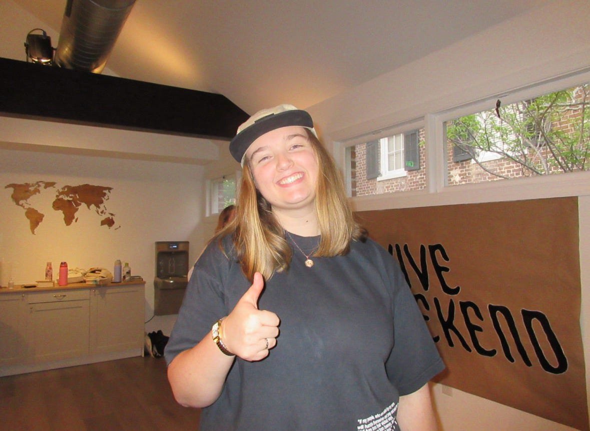 A young woman smiling and giving a thumbs-up gesture inside a room with a world map on the wall, a water fountain, and a sign that reads "LOVE SICKLEPO".