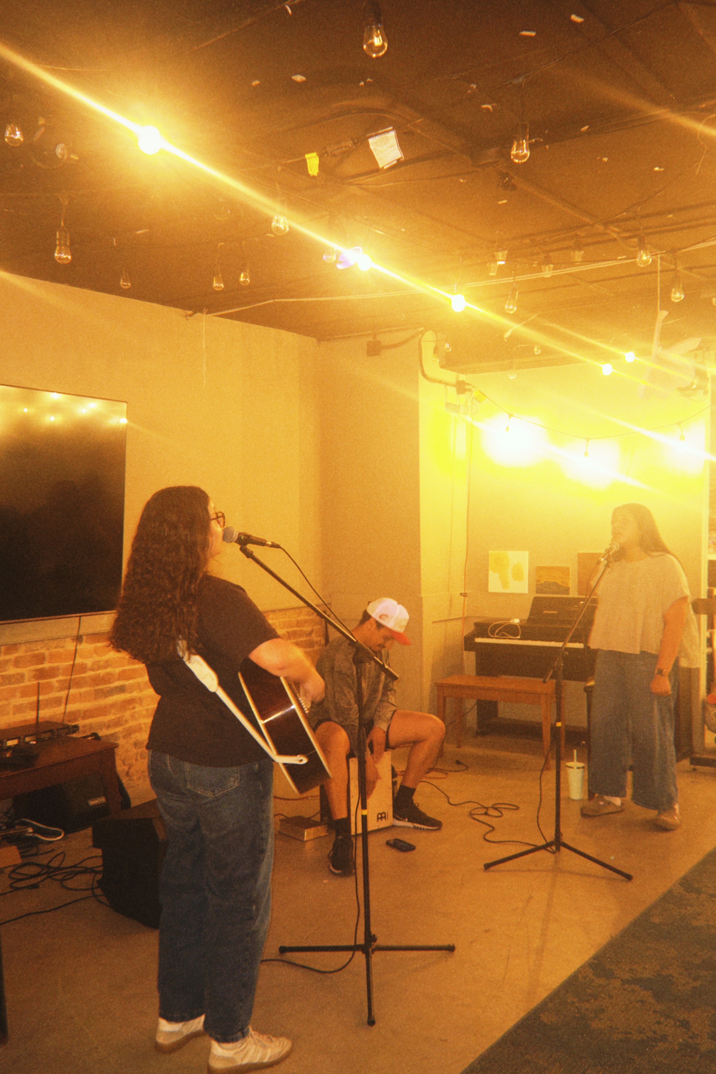 Four people performing in a cozy indoor music venue with string lights and a brick wall. One person is playing a guitar, another is sitting with a cajón, and two others are standing with microphones, one near an electric keyboard.