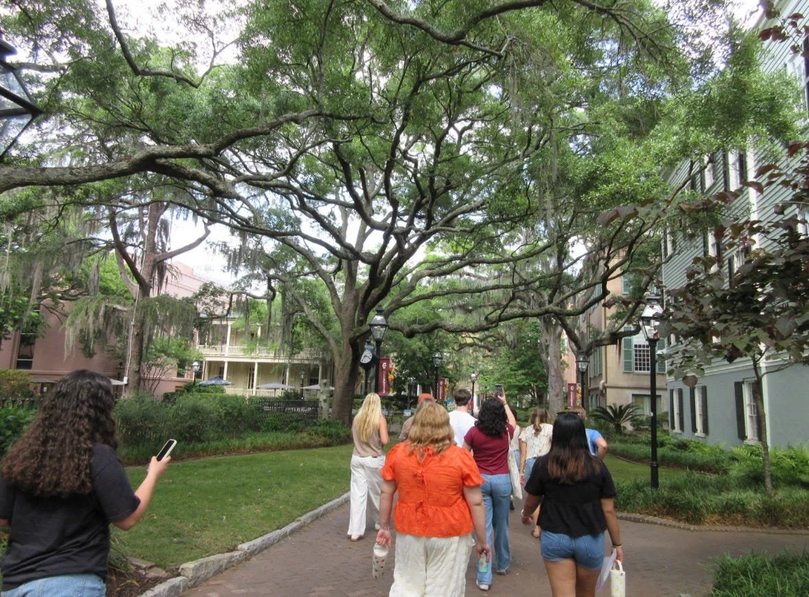 People walking along a path in a park with large trees and residential buildings in the background.