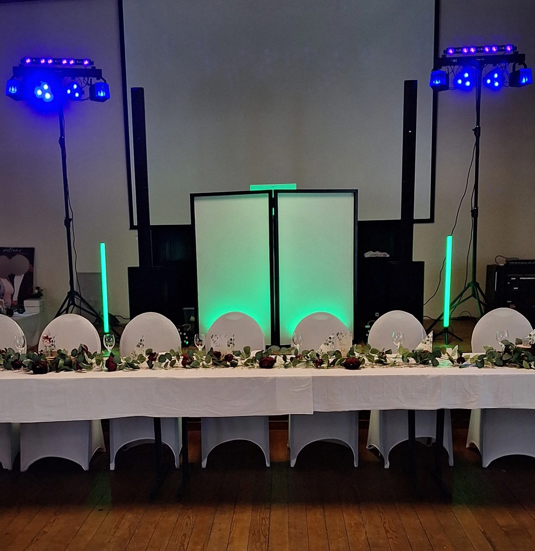 Der obere Teil zeigt eine Tanzfläche mit blauen Lichtstrahlern. Darunter befindet sich eine lange Tafel mit weißen Stuhlen, die mit Geschirr und Dekorationen für eine Feier gedeckt ist. Das Ganze ist in einem Raum mit Holzboden.