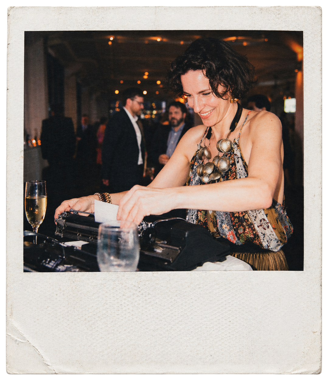 A woman is smiling while writing poems on a typewriter at a corporate social event with people mingling in the background.