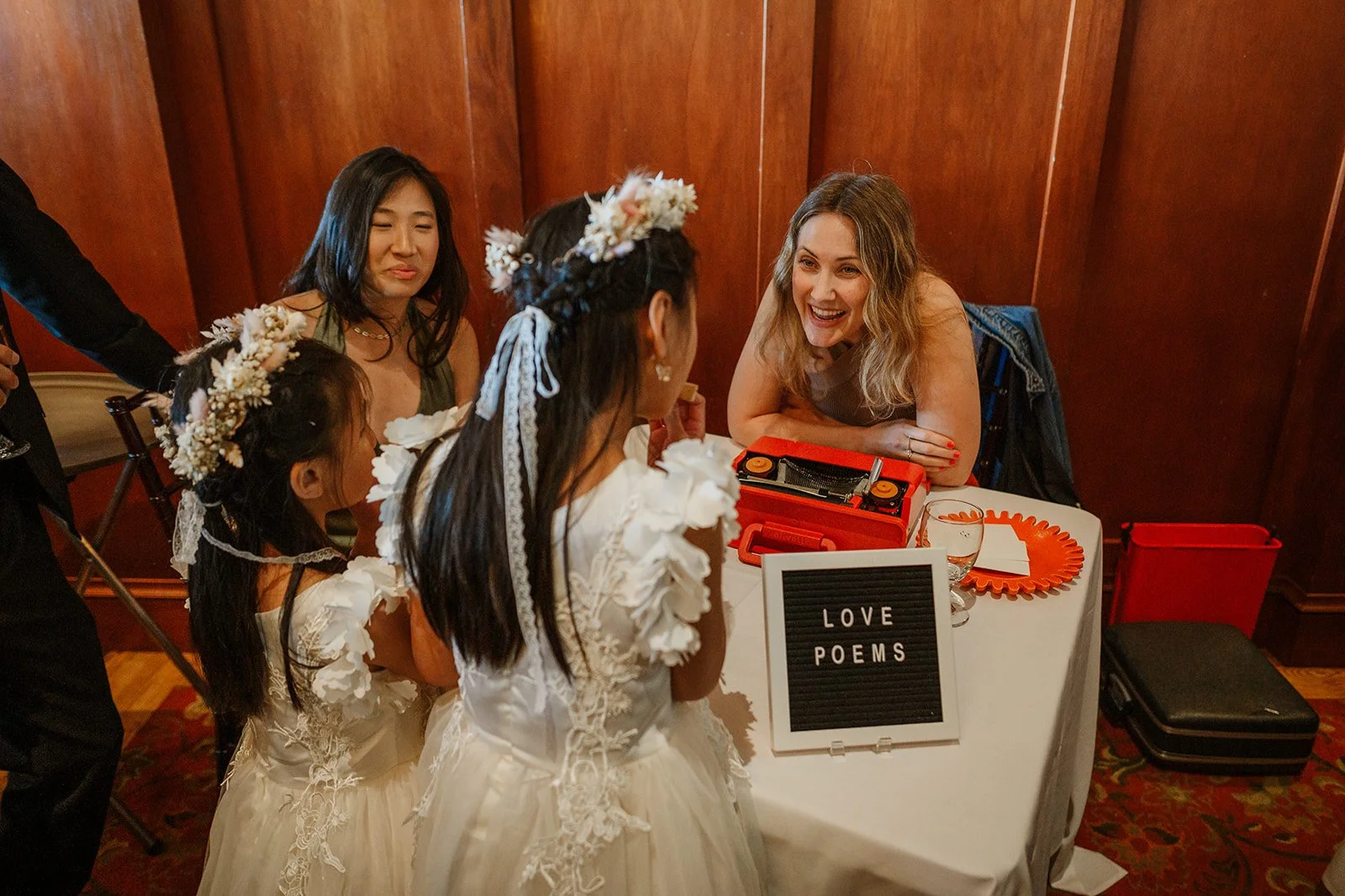 A woman with blonde curly hair sitting at a table talking to three girls dressed in white floral dresses with flower crowns, at a wedding or event getting Love Poems.