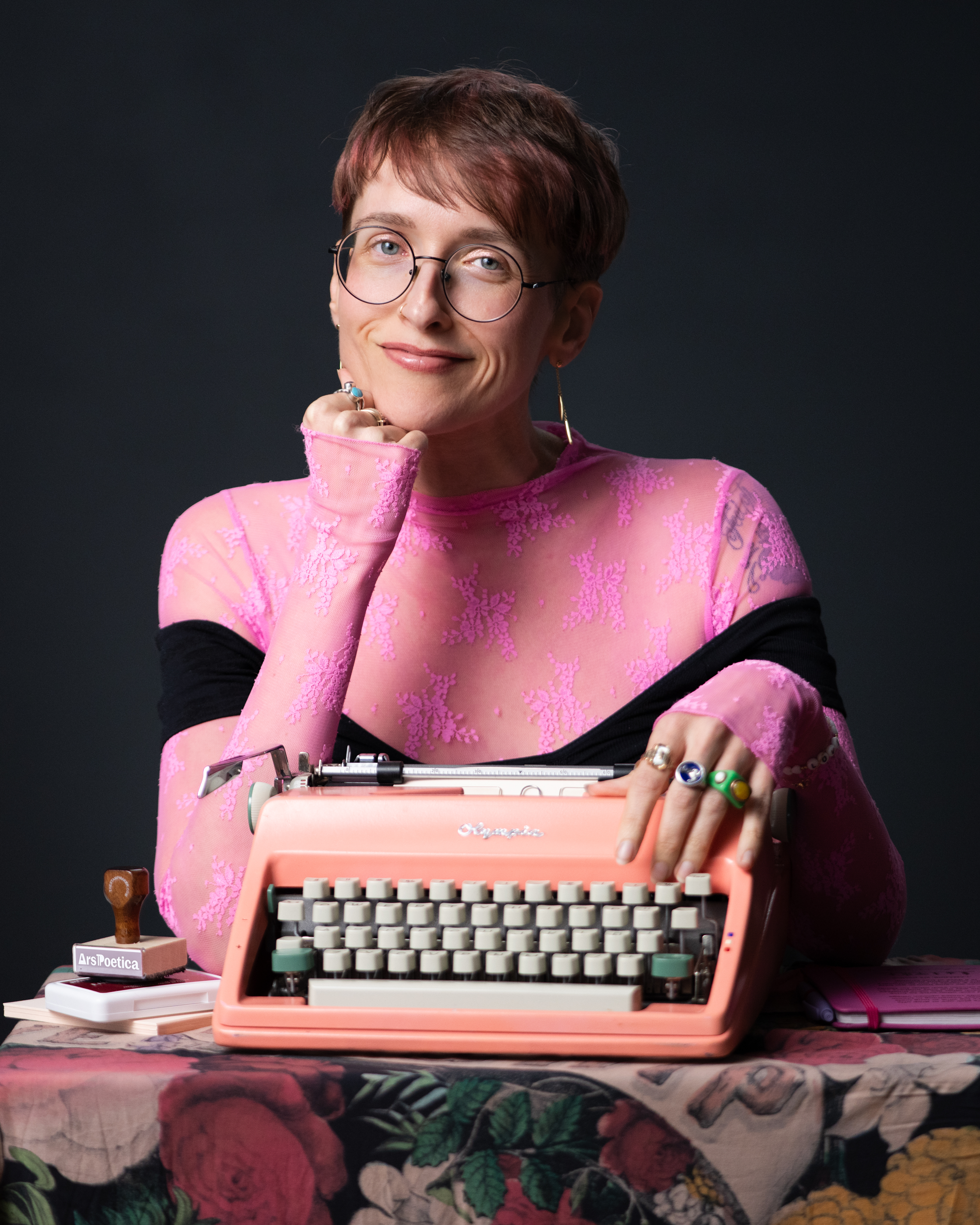 Person with short hair and glasses sitting at a table with an orange vintage typewriter, wearing a pink lace top and earrings, with books and a small sign on the table.