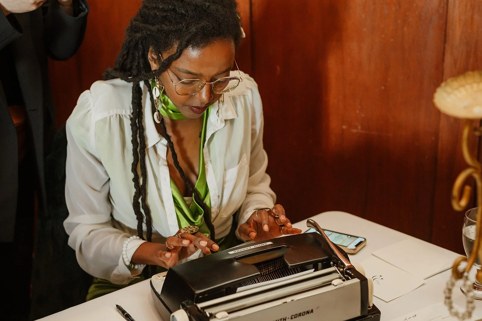A woman with glasses and dreadlocks sitting at a table, working on a document with a row of heated rollers in front of her.