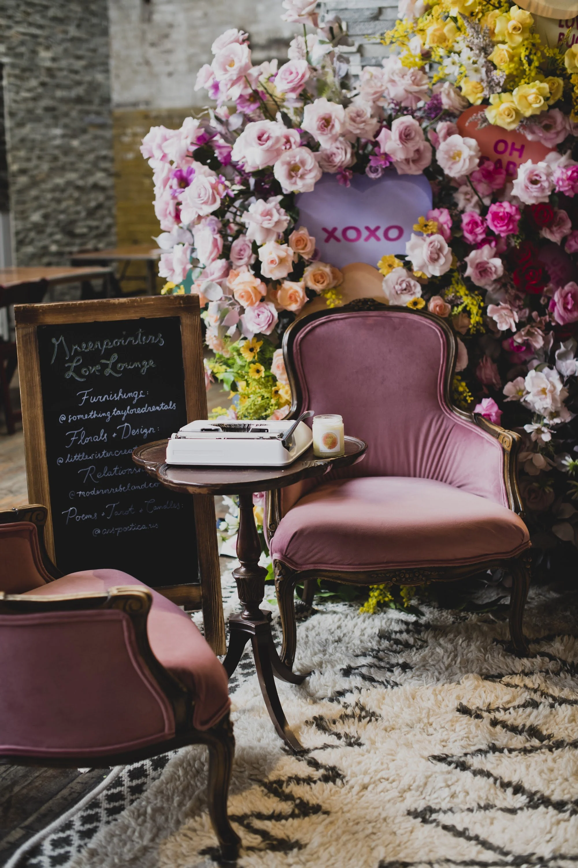 A cozy seating area with pink velvet chairs, a small round wooden table with a typewriter and candle, a large floral display with pink roses and yellow flowers, and a black chalkboard sign advertising a love lounge, all on a fluffy white rug.