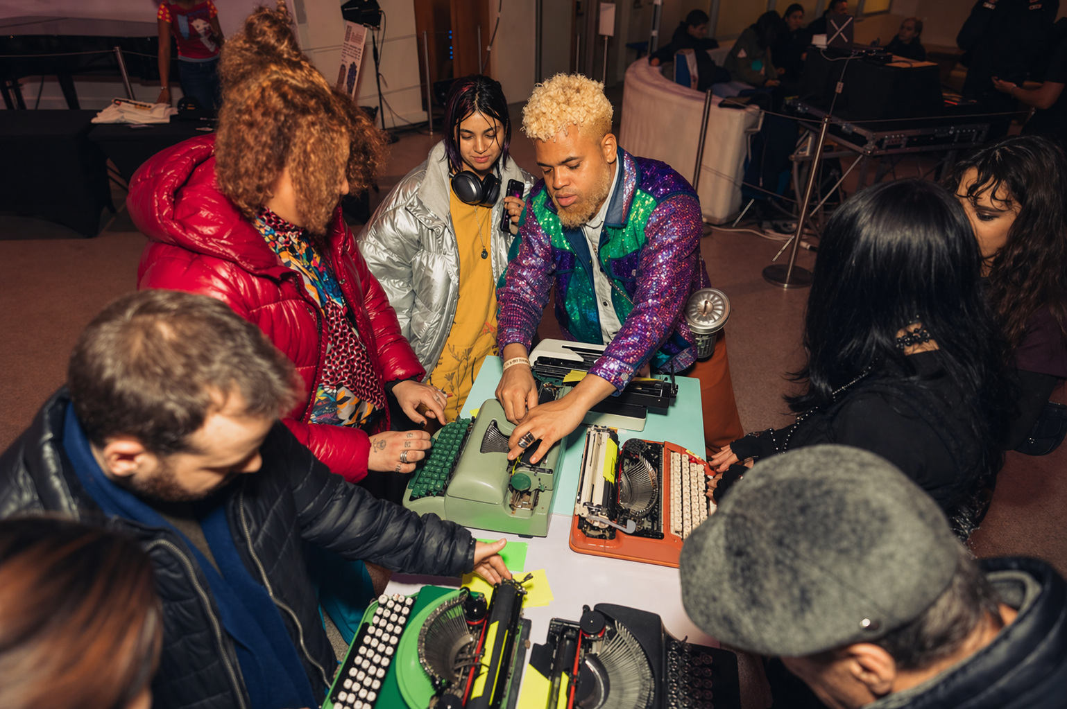 Group of people gathered around vintage typewriters on a table at an indoor event, with some individuals observing and others engaging with the typewriters.