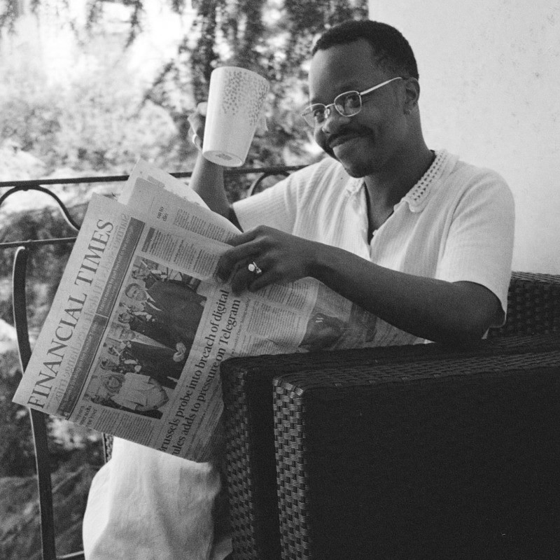 A man with glasses and a mustache reading a newspaper called the Financial Times, sitting outdoors on a balcony or porch, holding a mug, with trees and natural scenery in the background.