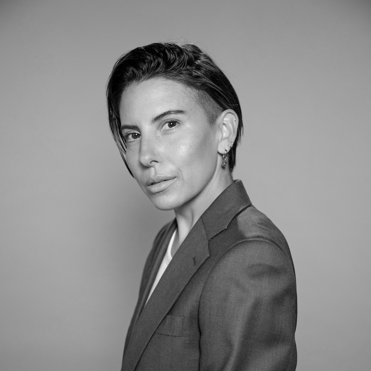 Black and white portrait of a person with short, styled hair, wearing a blazer and earrings, looking at the camera against a plain background.