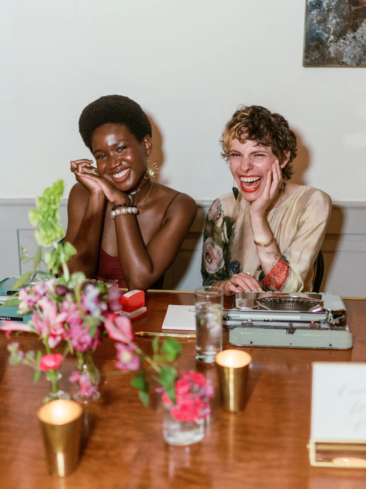 Two women sitting at a wooden table, smiling and laughing, with flowers, candles, a typewriter, and a glass of water in the foreground.