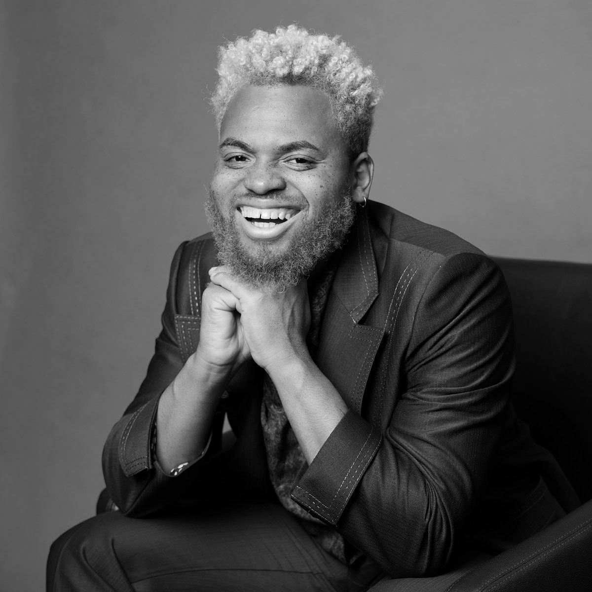 Black and white portrait of a man with curly blonde hair, beard, and earrings, smiling while sitting with his hands under his chin, wearing a dark blazer, against a plain background.