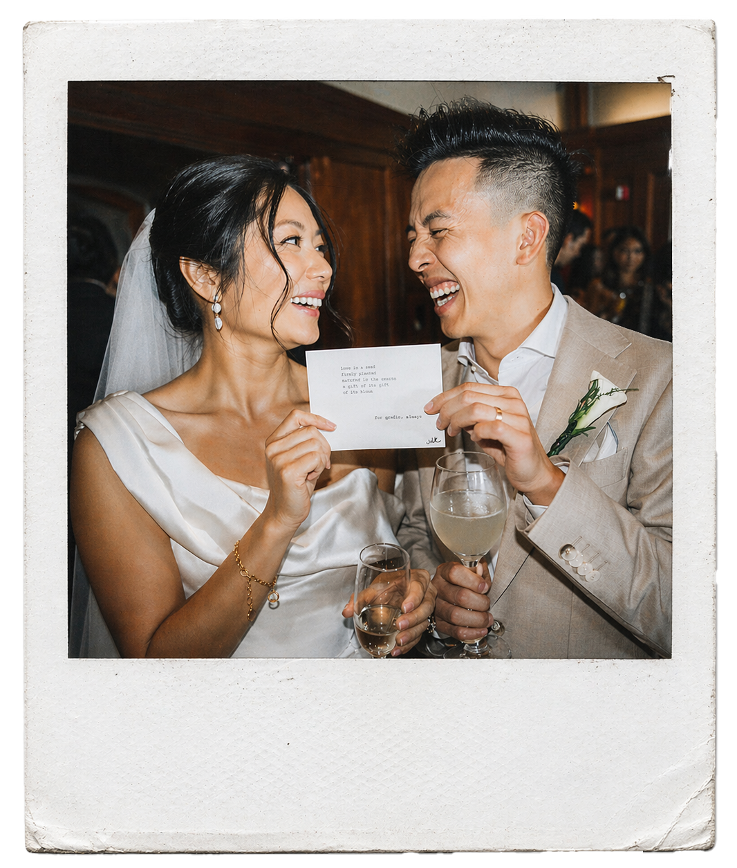 A bride and groom at their wedding celebration, smiling and holding a card with a poem on it together with champagne glasses in hand, in a warmly lit room with wooden walls in the background.