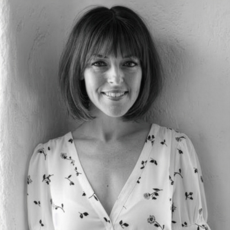 Black and white photo of a woman with shoulder-length hair, smiling, standing against a plain wall, wearing a floral patterned blouse.