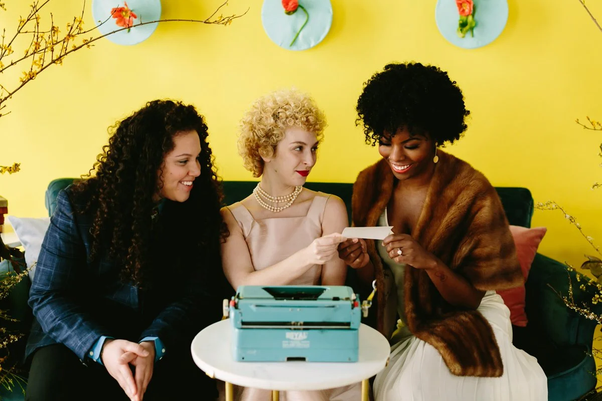 Three women sit on a green couch against a yellow wall decorated with paper plates and flowers. They are smiling and looking at a piece of paper, with a vintage typewriter on a white table in front of them.