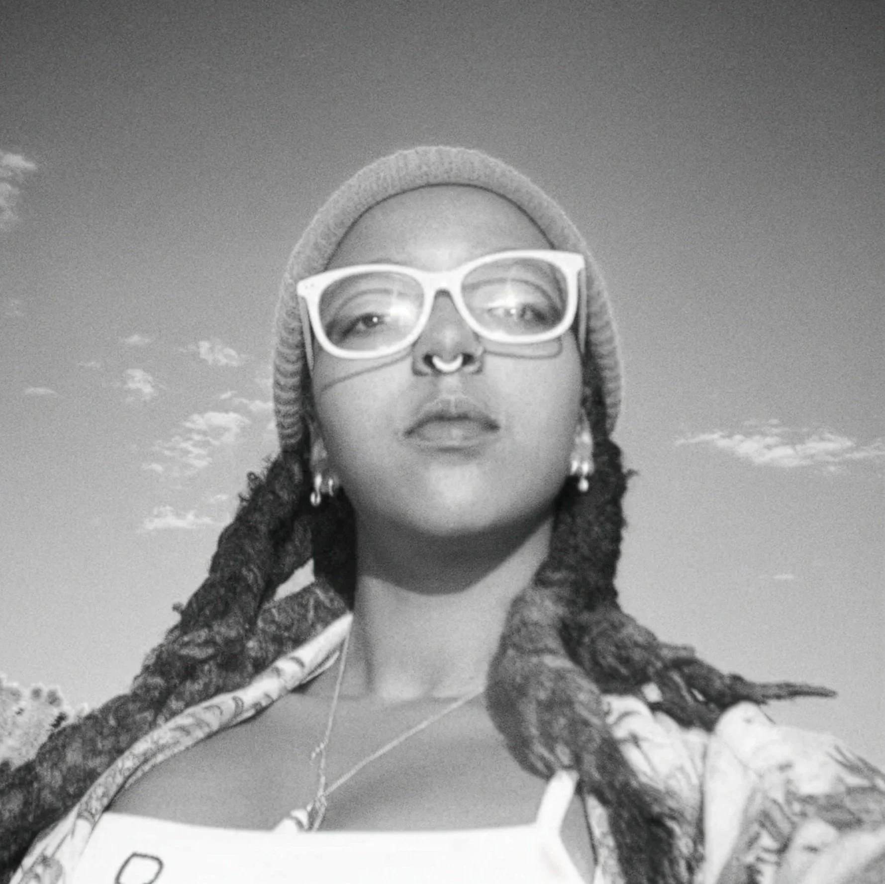 Close-up black-and-white photo of a person wearing glasses, a beanie, and a coat, with a cloudy sky in the background.