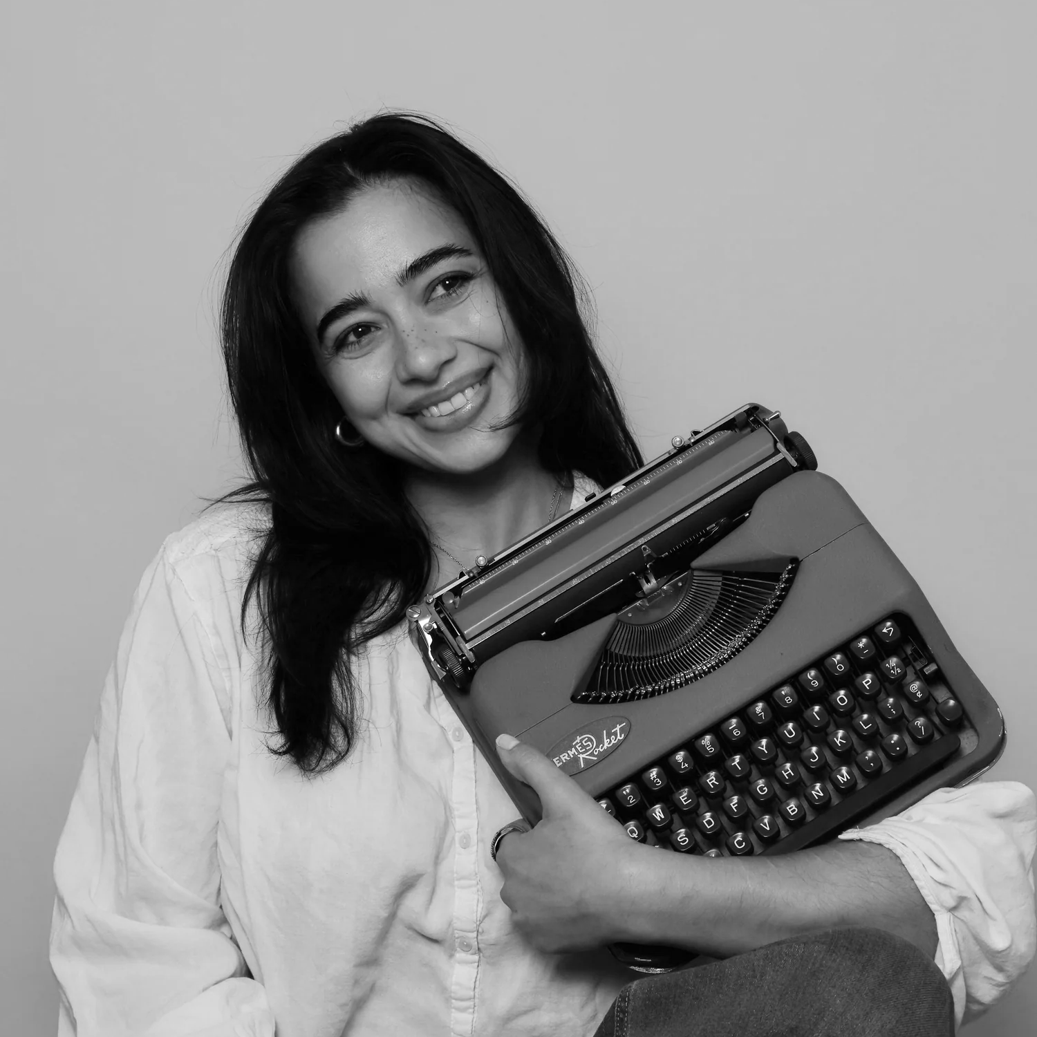 A young woman with dark hair, smiling, holding a vintage typewriter labeled 'Hermes Rocket', wearing a light-colored shirt.