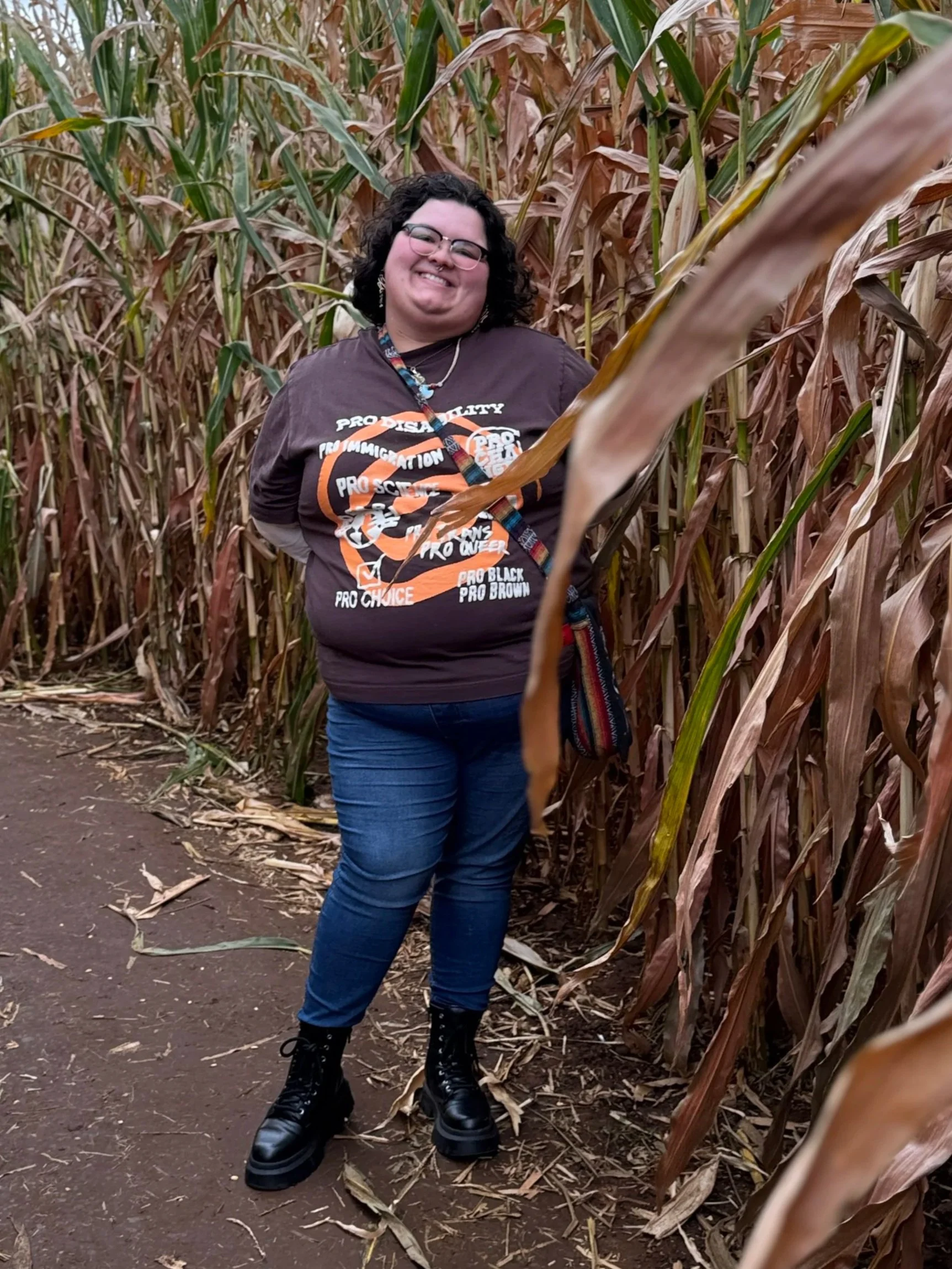 A woman with curly black hair, glasses, and earrings standing on a dirt path surrounded by tall, dried corn stalks. She is smiling, wearing a dark brown t-shirt with various slogans, blue jeans, and black boots, with a colorful strap across her chest.