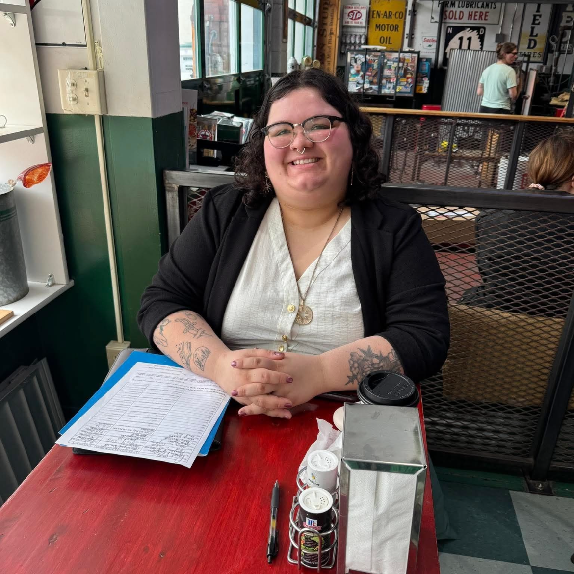 A woman with dark curly hair, glasses, and tattoos on her arms, smiling and sitting at a red table in a cafe or diner.
