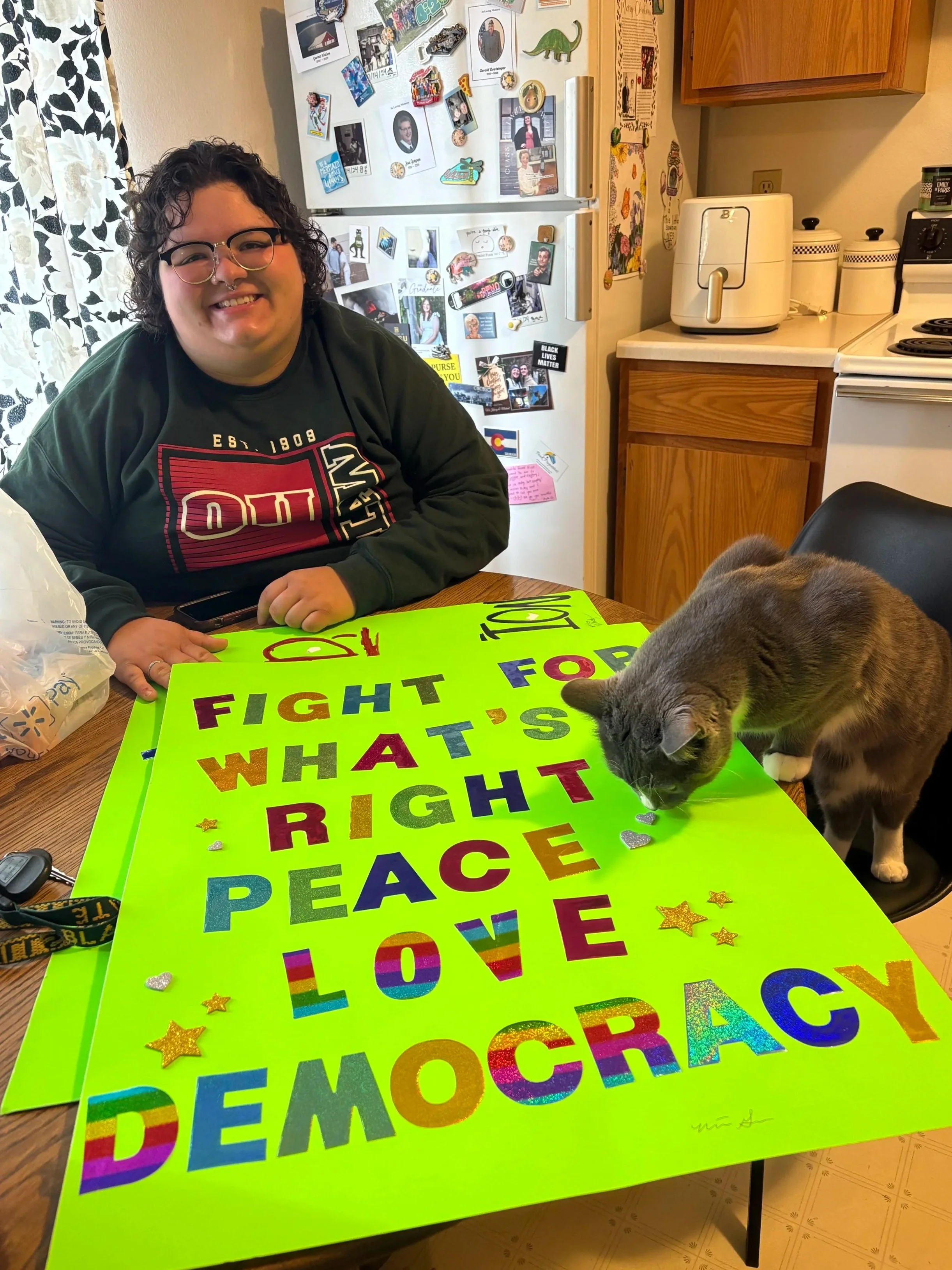A person sitting at a kitchen table smiling, with a cat sniffing a bright green poster that has colorful text promoting peace, love, and democracy. The background shows a refrigerator covered with photos and magnets, a wooden cabinet, and kitchen appliances.