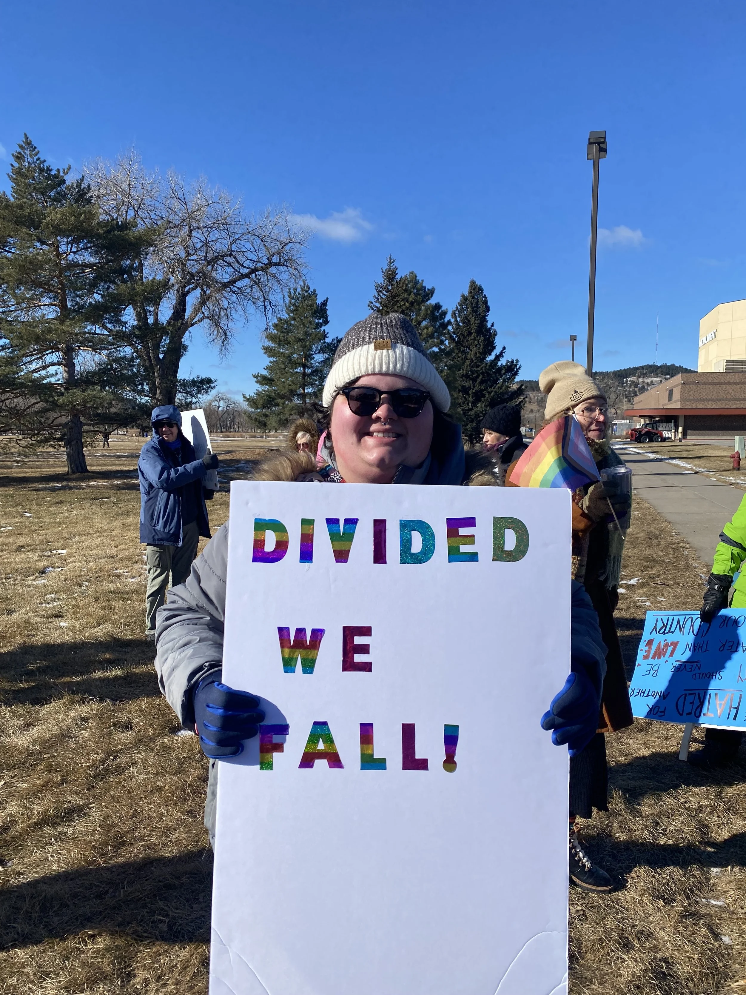 A smiling person wearing sunglasses, a beanie, and gloves holding a sign with colorful letters saying 'DIVIDED WE FALL' during an outdoor gathering or protest on a sunny day.