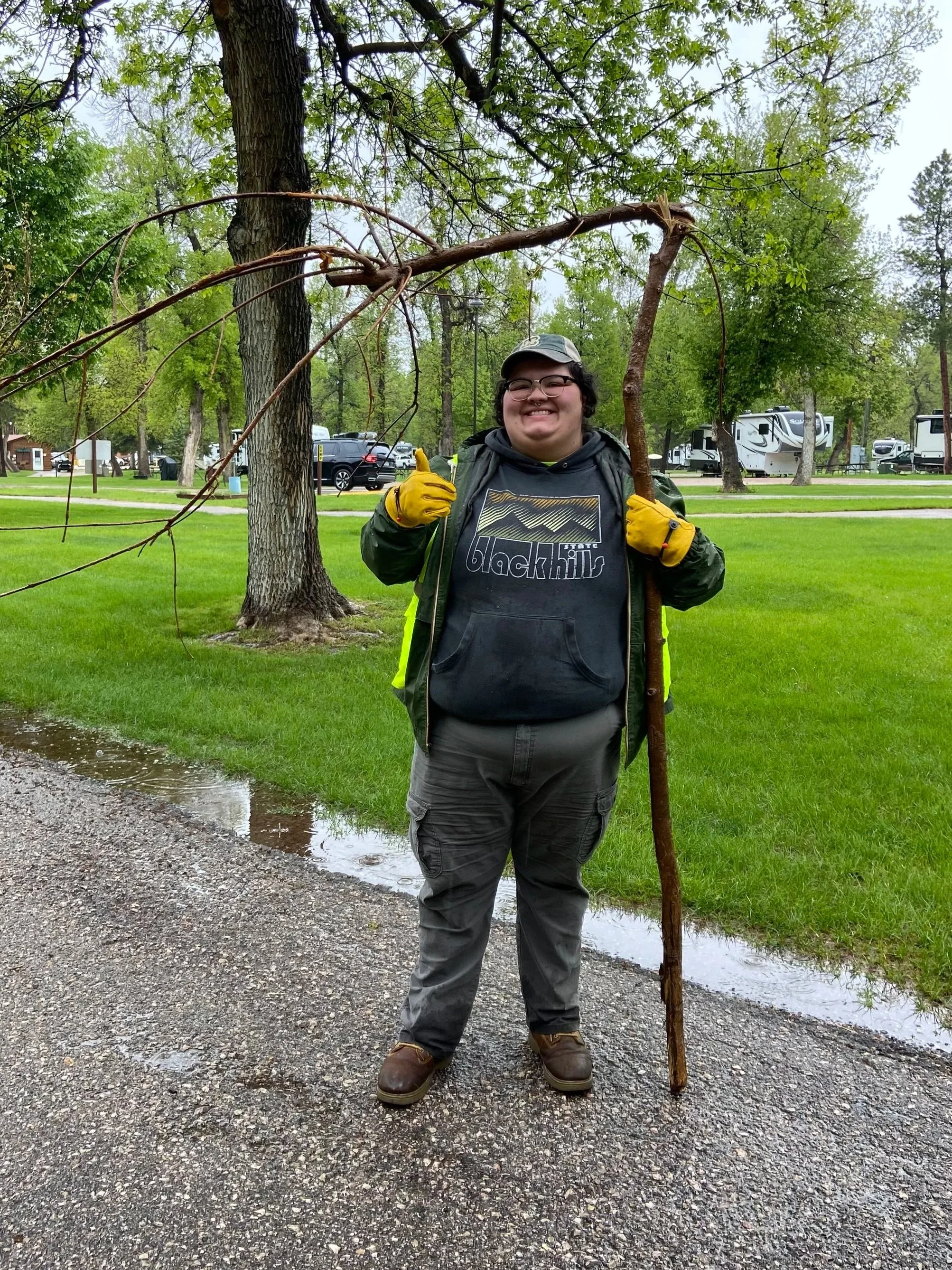 Person standing outdoors holding a large tree branch that resembles a stick and an arching vine, smiling at the camera, dressed in rain gear and yellow gloves, with a park and RVs in the background.