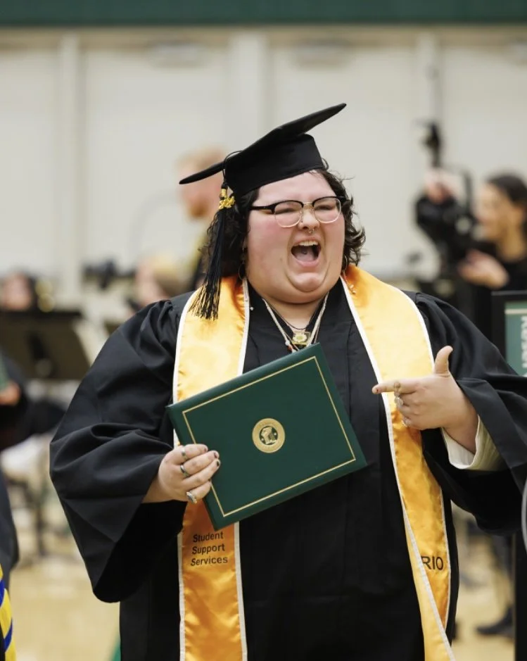 A graduate wearing a black cap and gown with a gold and white stole, holding a diploma, and smiling happily at a graduation ceremony.