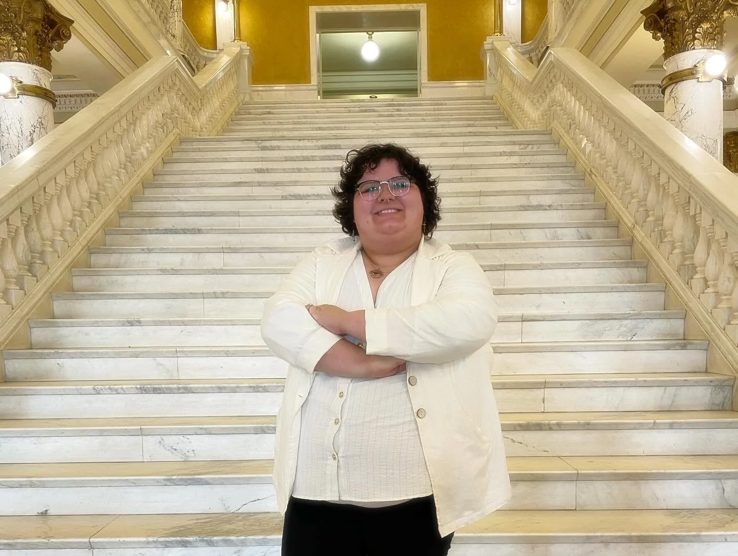 A person with short curly dark hair, glasses, and a septum piercing, standing with arms crossed in front of a grand marble staircase with ornate railings and a gold and marble interior background.