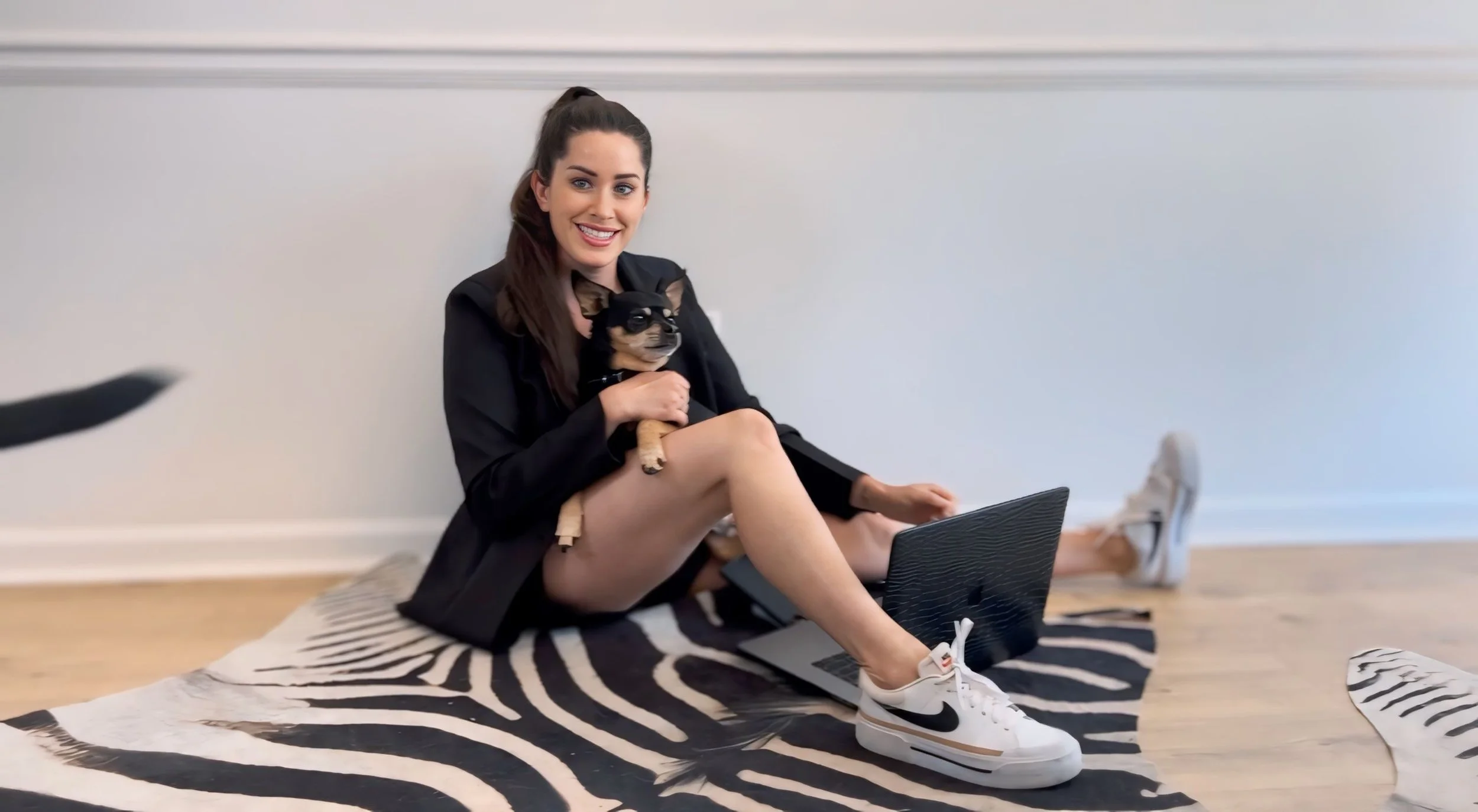 A young woman with long brown hair, wearing a black jacket and white Nike sneakers, is sitting on a zebra-striped rug on a wooden floor, holding a small black and tan Chihuahua dog, while a laptop is open in front of her.
