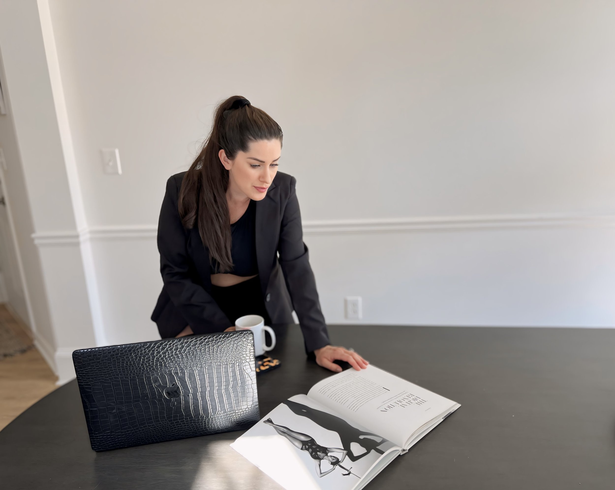 Woman in black blazer leaning over a dark table, looking at an open magazine with black and white images. A laptop with a crocodile skin pattern cover and a coffee mug are on the table.