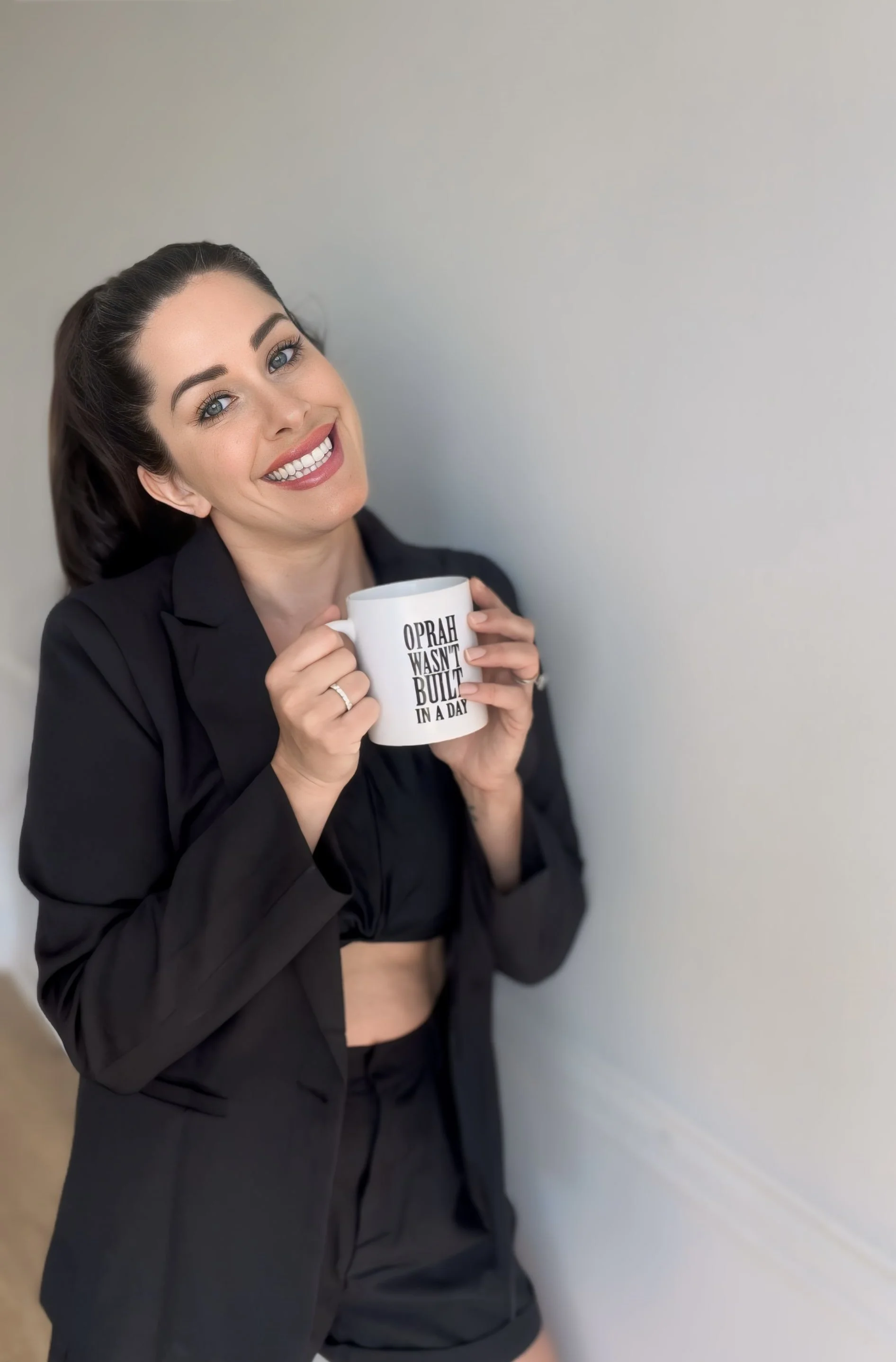 A woman with brown hair, smiling, wearing a black blazer and shorts, holding a white mug with the text 'Oprah wasn't built in a day', standing against a light-colored wall.