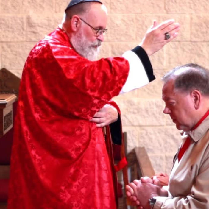 A priest wearing a red vestment performs a blessing over a man with gray hair, who is kneeling with his hands clasped in prayer.